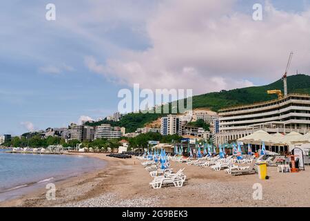 Des chaises longues avec parasols pliés se trouvent sur la plage, sur fond de bâtiments, de verdure et de montagnes Banque D'Images