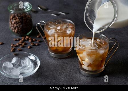 Deux tasses de café froid avec glaçons, dans lesquelles le lait est versé à partir d'un pot. Arrière-plan sombre. Gros plan Banque D'Images