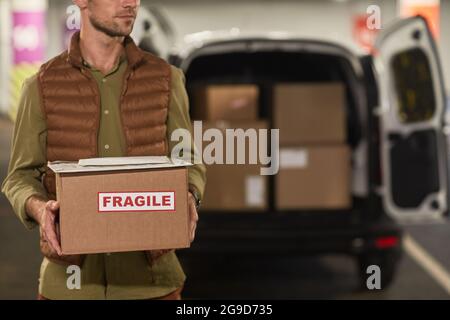 Portrait rogné d'un employé de livraison tenant des boîtes et souriant à l'appareil photo tout en se tenant près d'une camionnette à l'extérieur, espace de copie Banque D'Images