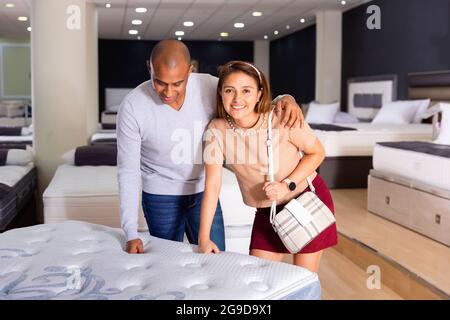 Jeune femme souriante latina avec son petit ami choisissant un nouveau matelas orthopédique moderne dans la boutique de meubles Banque D'Images