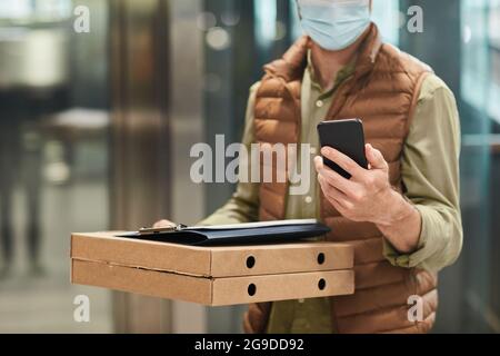 Portrait court de l'livreur portant un masque et tenant une pizza tout en utilisant le smartphone dans l'ascenseur, espace de copie Banque D'Images