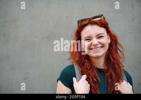Portrait d'une jeune femme en train de rire debout à l'extérieur sur fond gris, regardant l'appareil photo. Banque D'Images