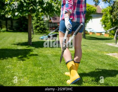 Femme jardinière marchant dans la cour tenant des sécateurs. Vue de derrière une jeune femme portant des bottes en caoutchouc et tenant des outils de jardinage. Banque D'Images