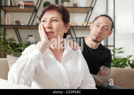 Triste mère mature assise sur un canapé et regardant loin avec son fils assis à l'arrière-plan et la soutenant Banque D'Images