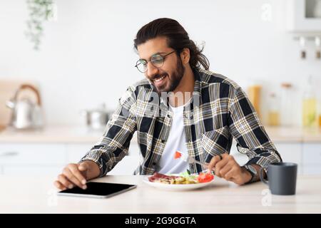 Joyeux garçon indien millénaire ayant le déjeuner et utilisant la tablette numérique, portant un ensemble décontracté et des lunettes, assis à la table de cuisine, manger frais sain s. Banque D'Images