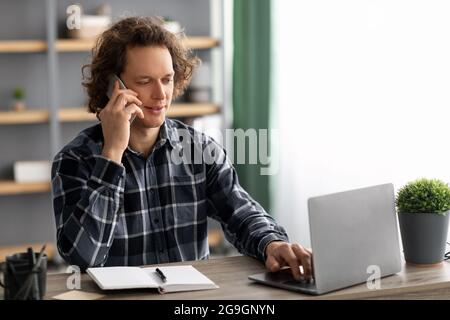 Vue latérale d'un homme d'affaires parlant au téléphone en utilisant un ordinateur portable assis sur un lieu de travail au bureau. Entrepreneur sérieux masculin ayant la compagnie de téléphone cellulaire Banque D'Images