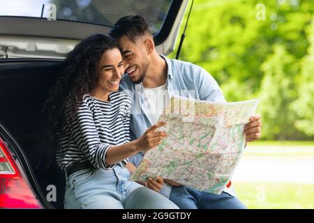 De jeunes amoureux heureux se mordillent en étant assis sur le coffre à bagages et en regardant la carte routière, l'espace de copie. Beau gars arabe liant avec sa belle petite amie, ha Banque D'Images