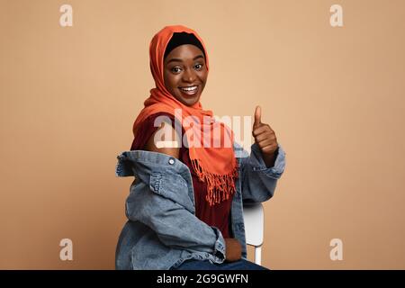 Belle jeune femme musulmane afro-américaine dans le hijab s'est vaccinée, femme noire souriante montrant l'épaule avec le bandeau médical et le pouce vers le haut, beige St Banque D'Images