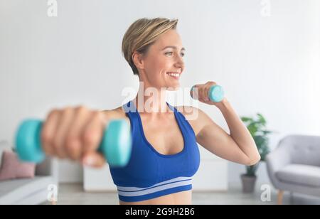 Concept de fitness domestique. Portrait d'une femme adulte souriante dans un soutien-gorge de sport s'exerçant avec deux haltères se posant à la maison, en appréciant l'entraînement, en regardant loin. Banque D'Images