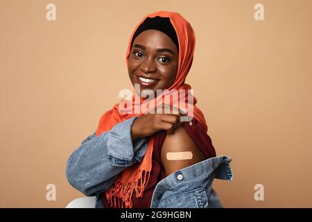 Une jeune femme afro-américaine joyeuse, musulmane, dans le hijab, s'est vaccinée, portrait en gros plan. Femme religieuse noire montrant l'épaule avec bande médicale, beige Banque D'Images