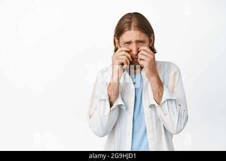 Portrait de mec blond triste et stupide, homme adulte pleurant et regardant bouleversé, essuyant les larmes et les yeux de frottement déprimé, debout contre le blanc Banque D'Images