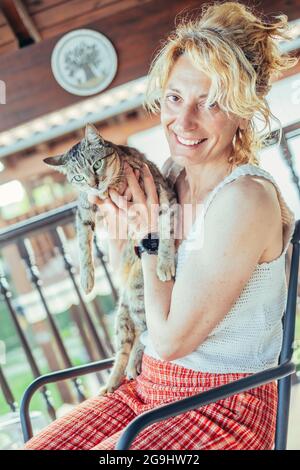 portrait d'une jeune femme blonde caucasienne mûre souriant avec son chat à la maison sur le porche assis dans une chaise à bascule. Concept de style de vie. Banque D'Images