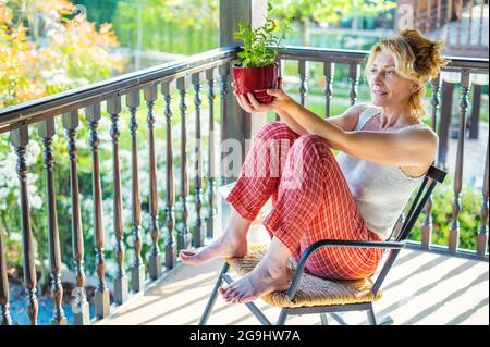 portrait d'une jeune femme blonde caucasienne mature se détendant à la maison sur le porche assis dans une chaise à bascule avec une plante en pot. Concept de style de vie. Banque D'Images