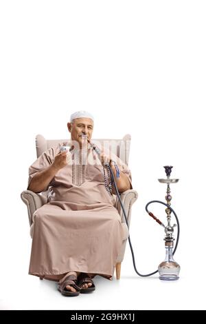 Homme mature dans des vêtements ethniques assis dans un fauteuil et fuyant shisha isolé sur fond blanc Banque D'Images