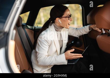 Bonne femme d'affaires parlant avec le conducteur et travaillant avec un ordinateur portable en étant assise dans la voiture Banque D'Images
