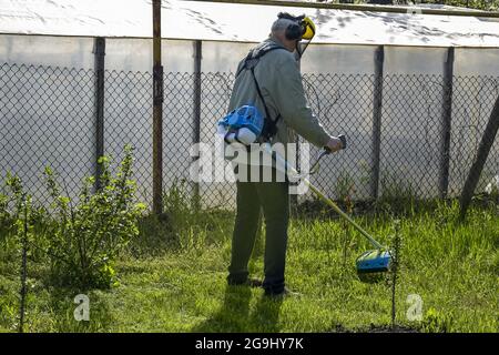 Le jardinier s'occupe du jardin. Processus de coupe de l'herbe verte avec le coupe-herbe dans le jardin. Port de protections auditives et visuelles. Tondeuse à essence. Banque D'Images