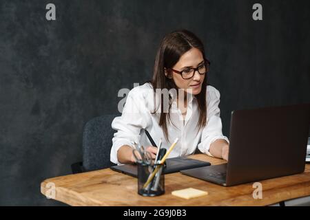 Femme de brunette pensive d'âge moyen travaillant sur ordinateur portable sur fond de mur sombre Banque D'Images