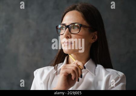 Femme de brunette pensive d'âge moyen en chemise blanche tenant un crayon sur son visage regardant de côté sur fond de mur sombre Banque D'Images
