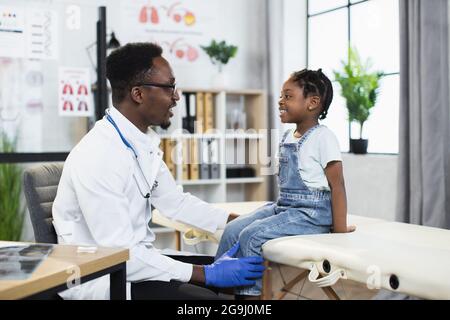 Vue latérale du médecin afro-américain en blouse de laboratoire blanche et gants examinant la santé de la petite fille mignonne à l'hôpital moderne. Concept de médecine et d'enfance. Banque D'Images