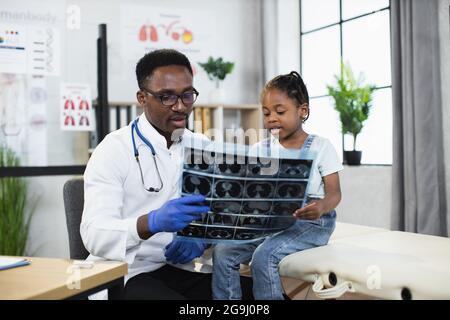 Médecin afro-américain en blouse de laboratoire blanche, lunettes et gants tenant un scanner à rayons X et expliquant les résultats à la petite fille. Concept des soins de santé et des personnes. Banque D'Images