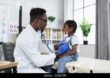 Vue latérale du pédiatre afro-américain utilisant le stéthoscope pour écouter les poumons de la petite fille à l'hôpital moderne. Médecin qualifié effectuant un contrôle de la patiente. Banque D'Images