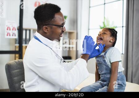 Jolie petite fille afro-américaine assise sur le canapé au bureau du médecin, tandis que le beau médecin noir examine sa gorge et sa bouche avec une spatule. Concept de pédiatrie Banque D'Images