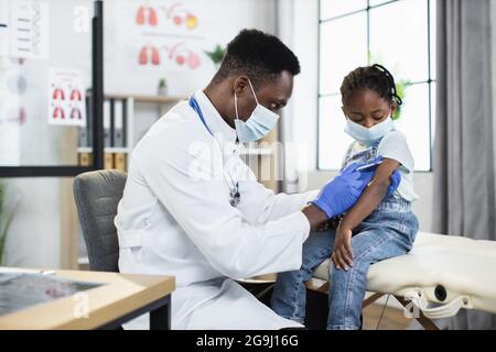 Médecin de sexe masculin utilisant une seringue pour vacciner une petite fille du coronavirus à l'hôpital moderne. Médecin afro-américain et patient portant un visage médical Banque D'Images