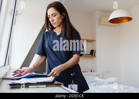 Une femme brune dans un costume médical bleu debout près de la fenêtre et lisant soigneusement les documents sur le fond de la pièce Banque D'Images