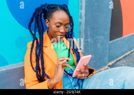 Italie, Milan, femme avec des tresses assis par un mur coloré, à l'aide d'un smartphone Banque D'Images