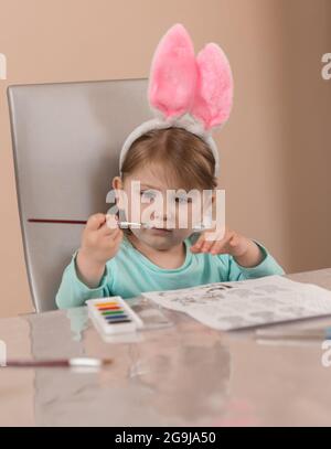 Une petite fille dans un joli bandeau avec des oreilles de lapin rose tire diligemment avec une brosse avec des peintures à l'aquarelle Banque D'Images