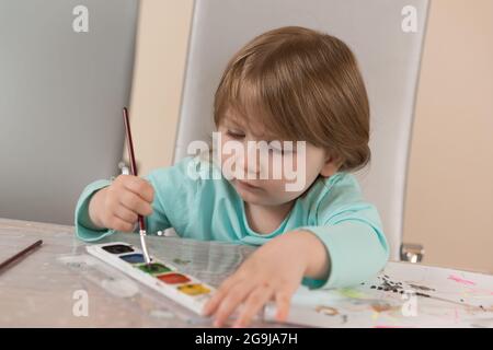 Une jeune fille de trois ans dans un chemisier turquoise à la table dessine avec des peintures à l'aquarelle Banque D'Images