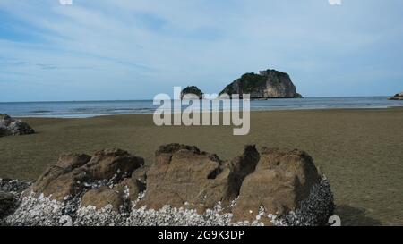 Huîtres sauvages attachées sur le récif rocheux à marée basse, vagues éclaboussant sur la plage de sable, la mer avec l'île en arrière-plan, parc national Khao Sam Roi Yot Banque D'Images