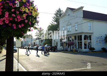 Scène de rue ensoleillée sur Bay Street dans la vieille ville de Florence, Oregon, avec des piétons, une architecture italienne historique et des fleurs suspendues colorées. Banque D'Images