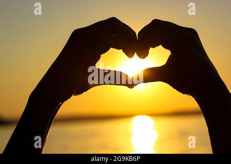 Silhouette des mains de la femme en forme de coeur au coucher du soleil contre un ciel jaune-orange, des rayons de soleil et un soleil à l'intérieur. Symbole de l'amour. Vacances par a s Banque D'Images