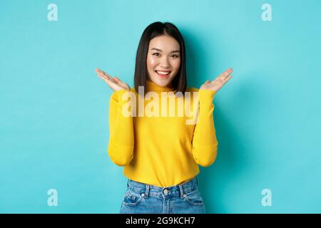 Beauté et mode. Une fille japonaise attirante lève les mains et montre quelque chose, souriant heureux et regardant l'appareil photo, montrant promo, blu Banque D'Images