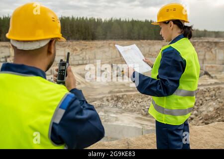 Femme ingénieur regardant le croquis et son collègue utilisant un talkie-walkie sur le chantier Banque D'Images