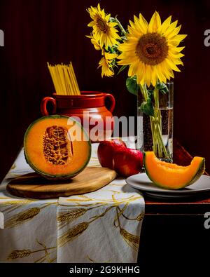 Un melon en deux et une tranche sur une assiette avec deux pommes rouges, des tournesols et des spaghetti crus, sur fond sombre Banque D'Images