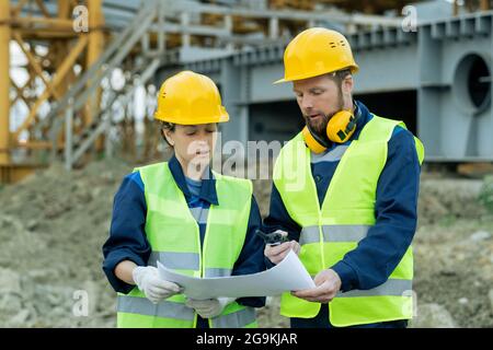 Deux ingénieurs en vêtements de travail et en casques de travail examinent le plan tout en travaillant en équipe sur le chantier de construction Banque D'Images