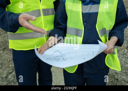 Gros plan de deux ingénieurs en vêtements réfléchissants examinant le plan d'un nouveau bâtiment à l'extérieur Banque D'Images