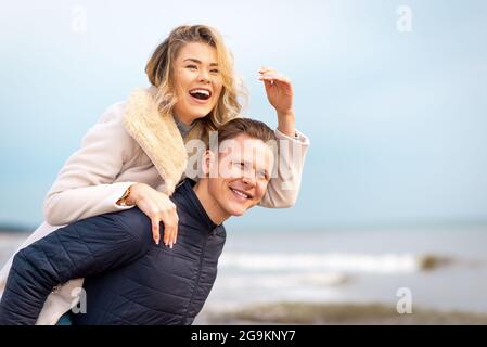 Joyeux homme qui donne le pigeyback à sa femme et riant à une plage. Souriant gars dans l'amour portant sur son dos sa petite amie et avoir plaisir.heureux en bonne santé Banque D'Images