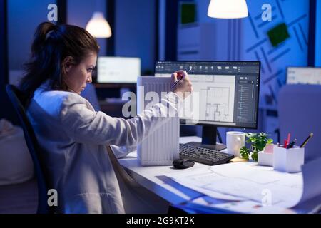 Femme architecte analysant et assortissant des plans pour le projet de nouveau bâtiment assis à un bureau. Graphiste utilisant le prototype arhitecture plans travailler des heures supplémentaires dans le bureau de l'entreprise tard la nuit Banque D'Images