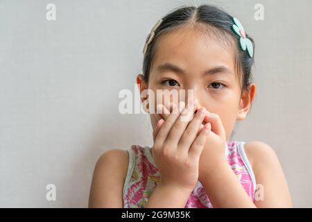 Portrait en gros plan de petite fille asiatique avec des cheveux longs noirs attachés, couvrant sa bouche avec deux mains. Banque D'Images