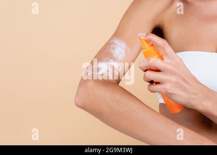 vue rognée d'une femme pulvérisant de la crème solaire sur le bras isolé sur beige Banque D'Images