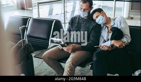 Un couple de touristes lassés s'est retrouvé coincé à l'aéroport pendant une pandémie. Homme utilisant un téléphone portable assis à côté d'une femme à l'aéroport. Les deux dans les masques faciaux. Banque D'Images