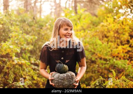 Portrait d'une belle jeune femme blonde heureuse avec de longs cheveux droits et un maquillage de vamp foncé regardant de côté, portant robe de guipure noire, tenant la citrouille Banque D'Images