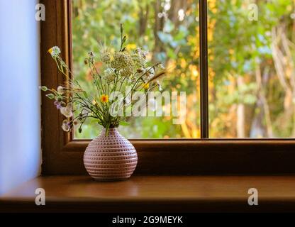 Bouquet de différentes fleurs sauvages dans un vase rose sur un rebord de fenêtre en bois contre une fenêtre en été ensoleillé. Fleurs à l'intérieur de la maison. Ambiance chaleureuse Banque D'Images