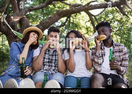 Souriant, les jeunes hommes et les femmes boivent de la bière et mangent des hamburgers pendant le pique-nique dans le jardin d'été. Quatre amis différents passent du temps libre ensemble sur l'air frais. Banque D'Images