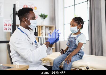 Pédiatre africain compétent dans le masque médical et les gants injectant le vaccin à la mignonne africaine fille. Médecin utilisant une seringue stérile pendant la procédure à l'hôpital. Banque D'Images