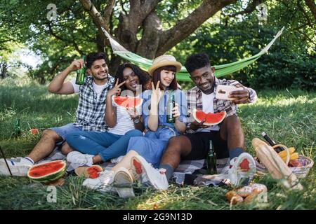Jeunes gens multiraciaux utilisant un smartphone moderne pour prendre le selfie dans un jardin verdoyant. Quatre amis joyeux s'amusant pendant un pique-nique dans la nature. Banque D'Images