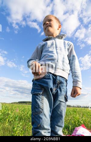 Un enfant d'âge préscolaire satisfait en jeans et un chandail tient l'argent de papier dans la nature dans un champ sur le fond d'un ciel bleu avec des nuages. Portrait Banque D'Images